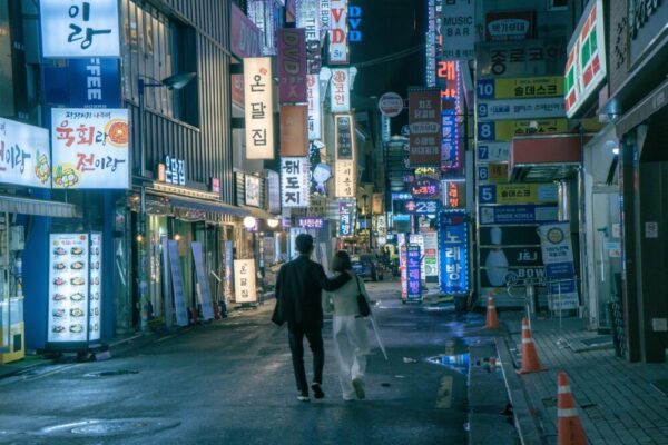 An Asian couple walks through the vibrant, neon-lit streets of Seoul at night, surrounded by shops and signs