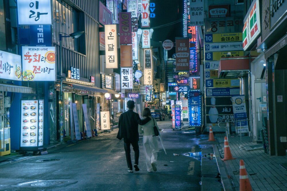 An Asian couple walks through the vibrant, neon-lit streets of Seoul at night, surrounded by shops and signs