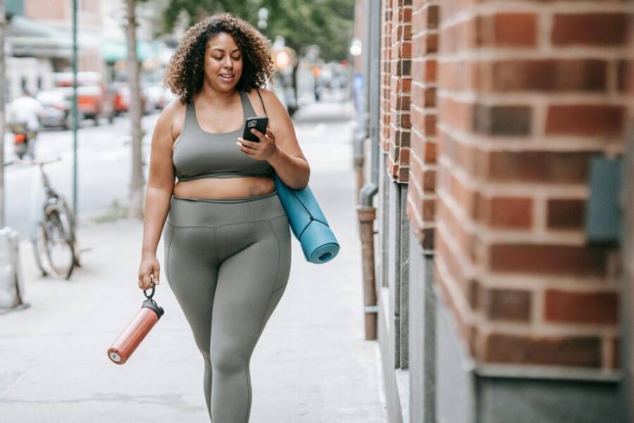 Confident woman in sportswear walking outside with yoga mat and phone, embracing positivity