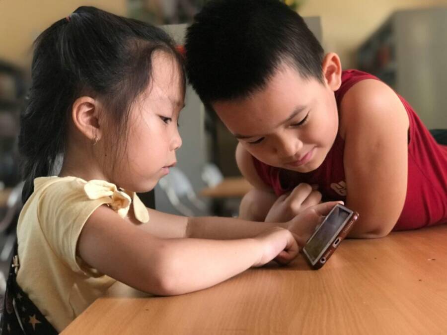 Young East Asian siblings intently watching a video on a smartphone indoors.