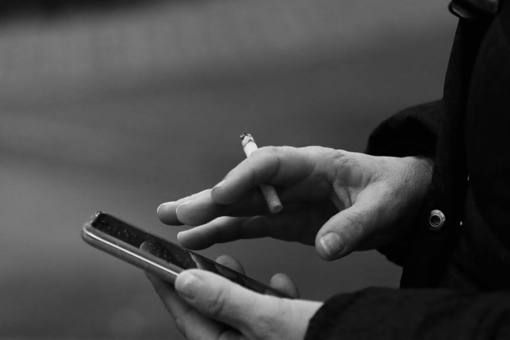 Monochrome image of hands texting on a smartphone with a cigarette, conveying technology and lifestyle