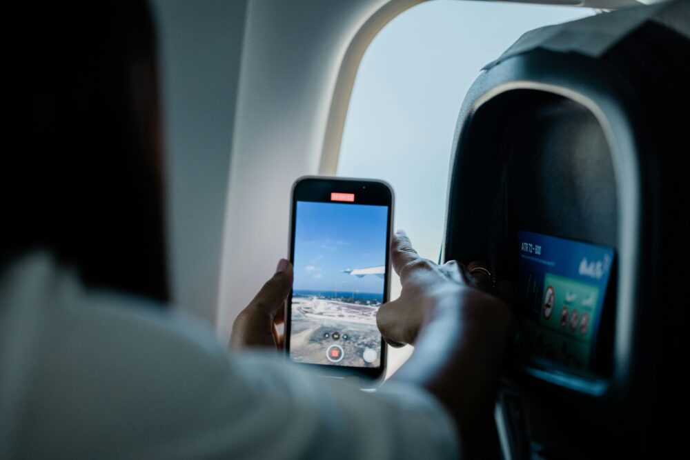A passenger films the outside view from an airplane window using their smartphone, capturing the wing and sky