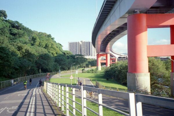 Cyclists ride under a vibrant viaduct near lush greenery and urban buildings in Taipei, Taiwan