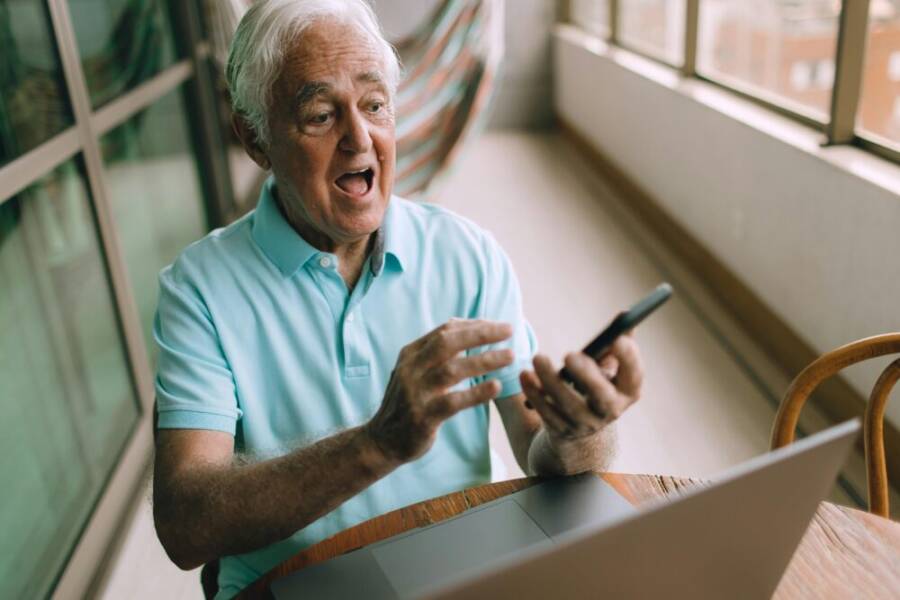 Elderly man in blue shirt reacts with surprise while using smartphone indoors.