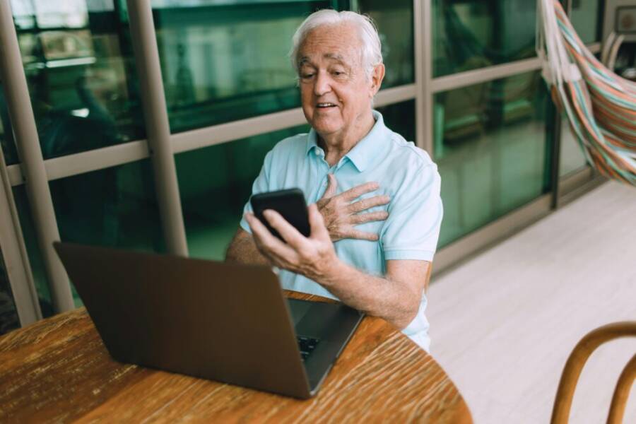 Elderly man using smartphone and laptop for video call at home, engaging with technology