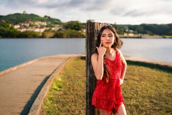 Young woman in a red dress stands by a lakeside, exuding confidence.