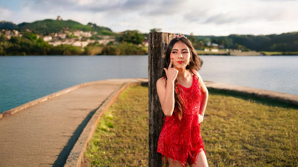 Young woman in a red dress stands by a lakeside, exuding confidence.
