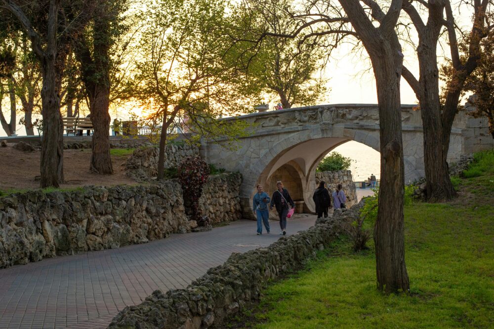 People enjoying a stroll under a picturesque stone bridge in a park at sunset with lush greenery.