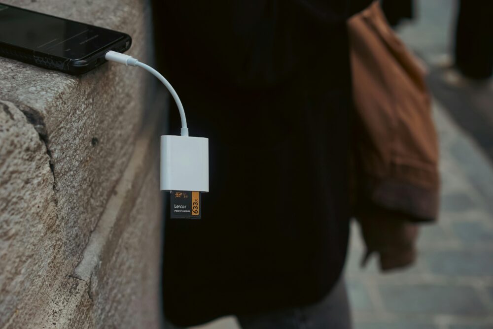 A mobile phone connected to a memory card reader outdoors on a stone surface.