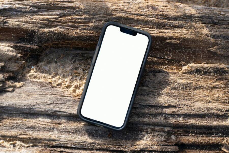 iPhone mockup with blank screen on a sandy wooden log at Kamala Beach, Phuket, Thailand.