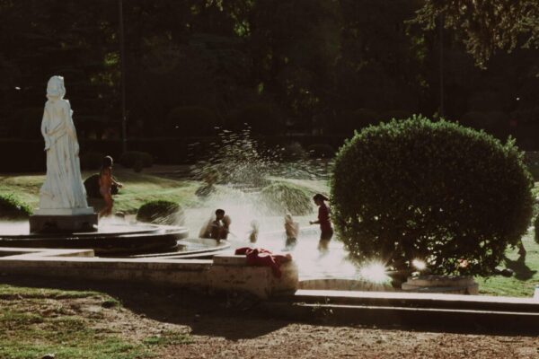 Children joyfully play in a fountain on a sunny day near a park statue.