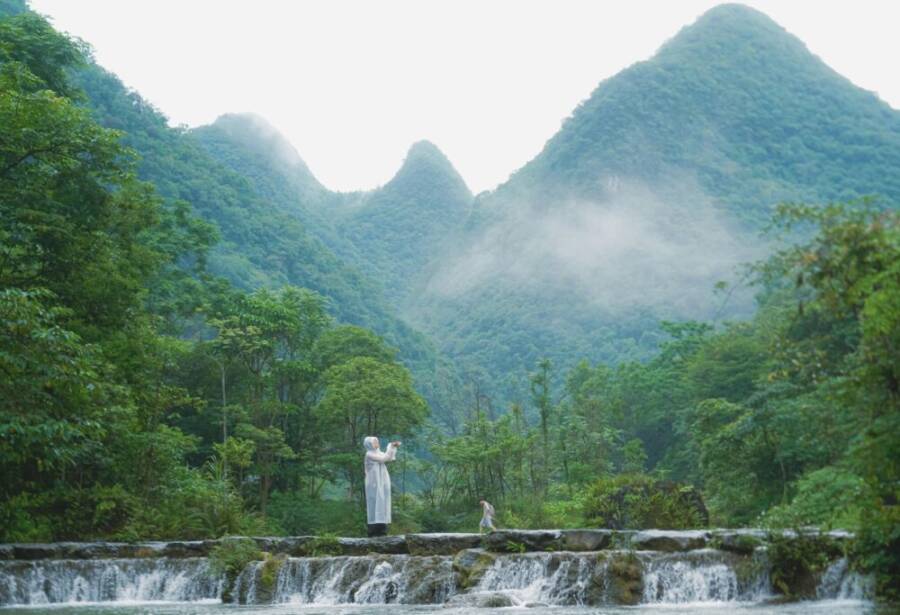 A peaceful scene of a person photographing a lush waterfall amidst misty mountains