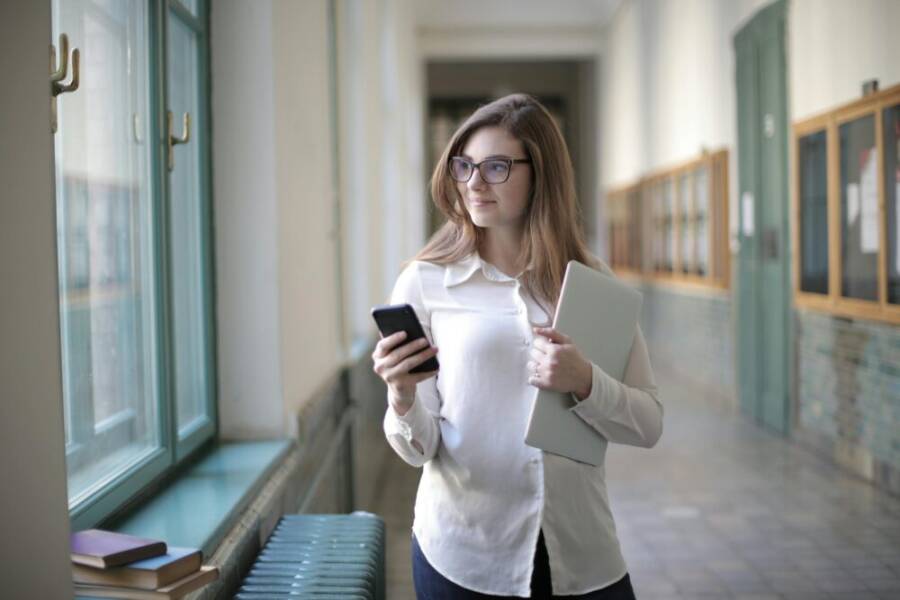 Female student with glasses holding a phone and laptop near window in school hallway