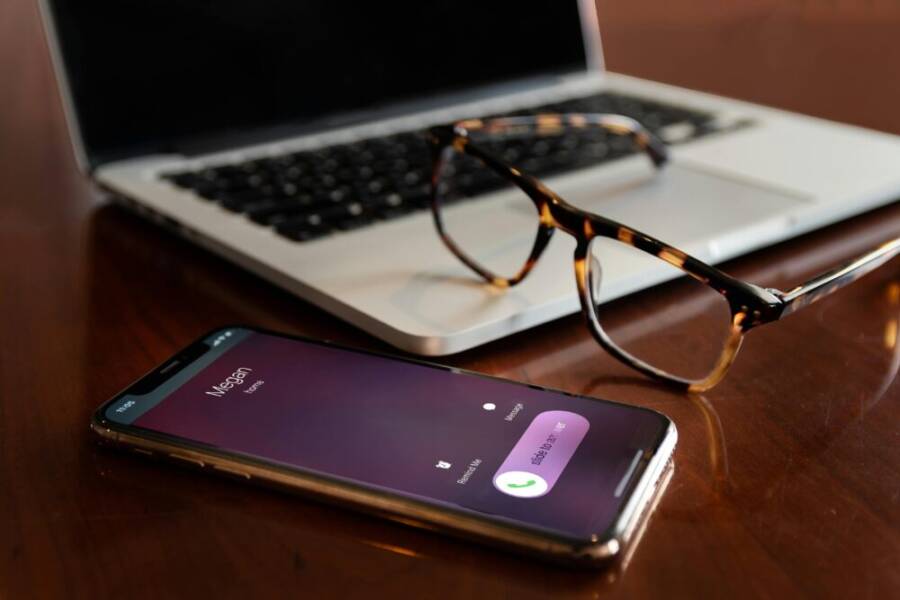 Close-up of a smartphone and laptop on a wooden desk with eyeglasses. Incoming call displayed.