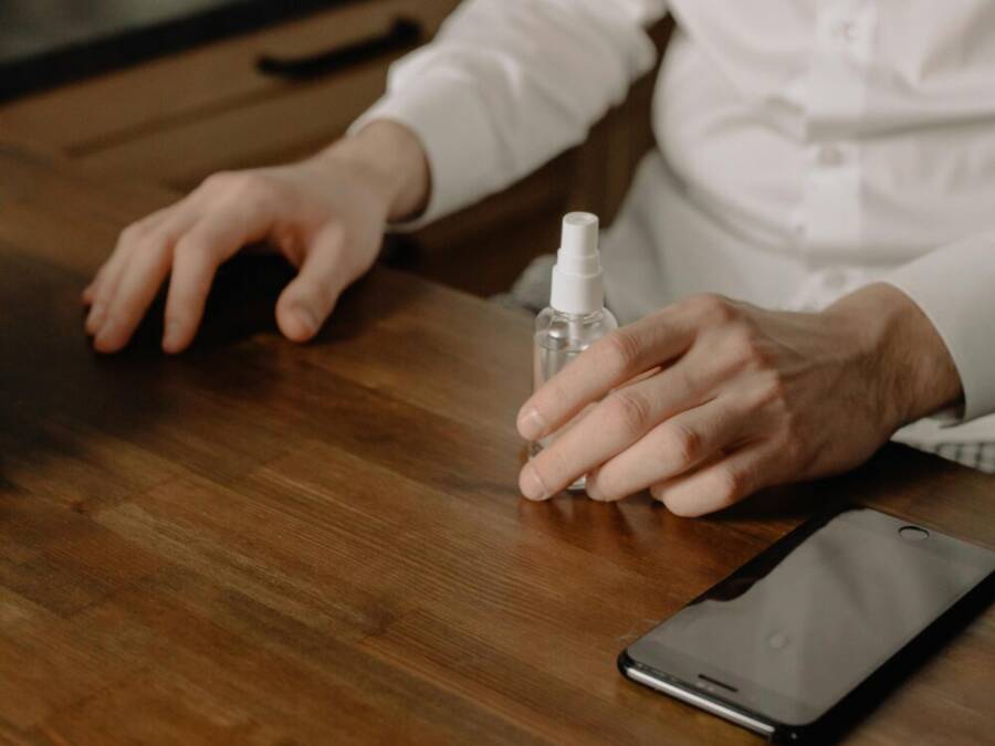 Close-up of hands using sanitizer spray beside a smartphone on a wooden table indoors