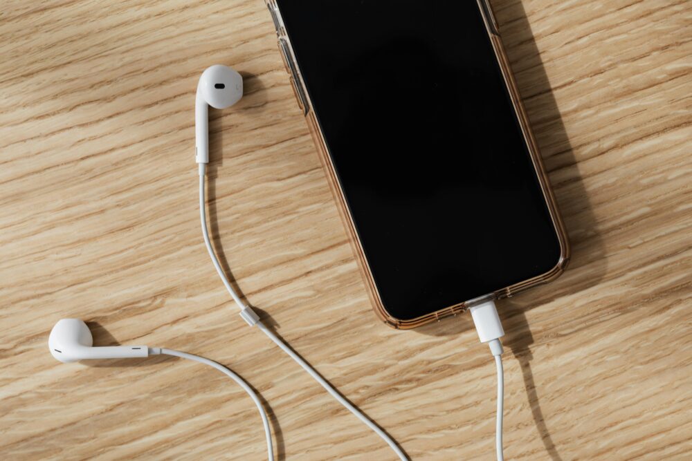 From above composition of white wired earphones and modern mobile phone with blank screen and connected charging cable placed on wooden surface in daylight