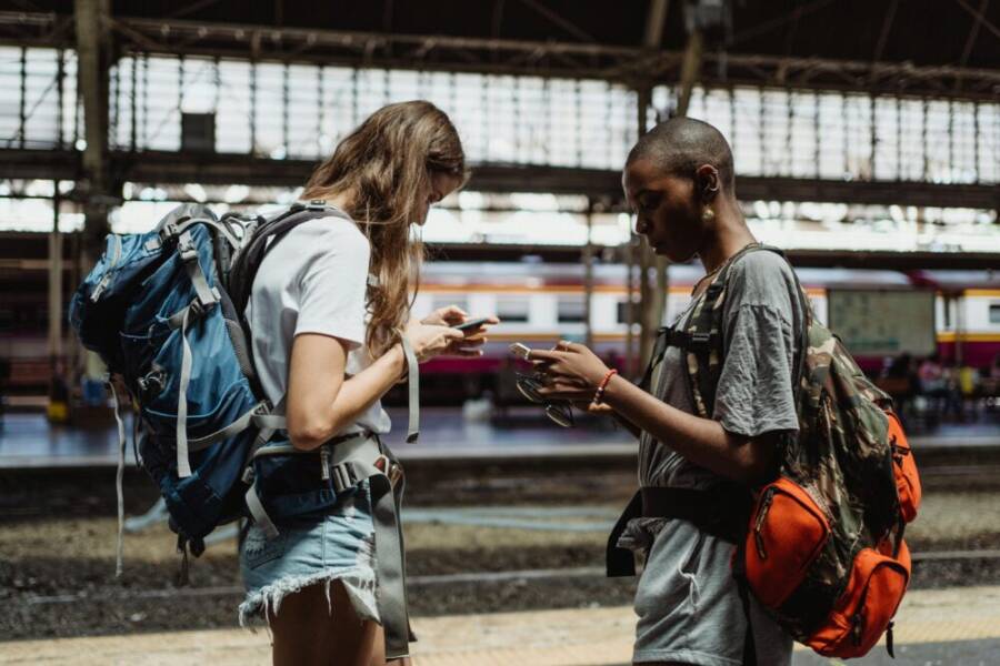 Two young women travelers using smartphones at a train station, planning their journey