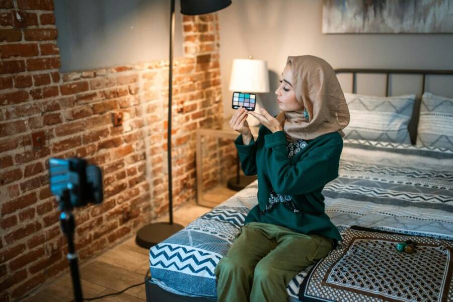 Muslim woman in hijab vlogging makeup tutorial with eyeshadow palette in bedroom setting