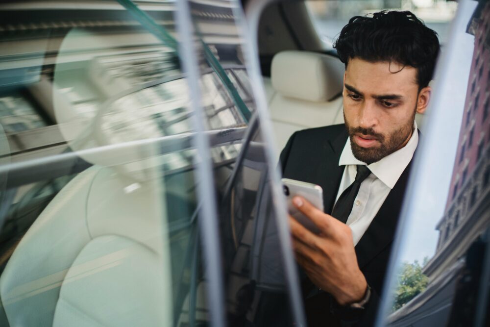 Confident businessman in suit using smartphone inside car, viewed through window reflection