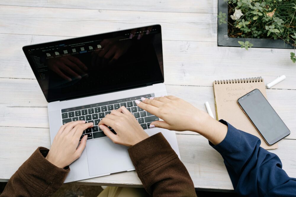 Two people collaborate on a laptop at an outdoor wooden table, with a smartphone and notebook nearby.