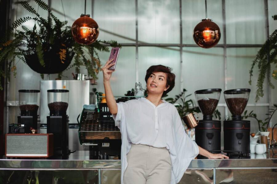 Cheerful Asian female with short brown hair in casual clothes standing against coffee machine and taking selfie on smartphone