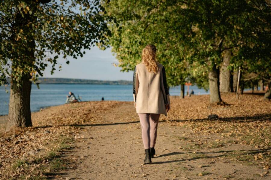 A woman walks along a tree-lined lakeside path in autumn sunshine