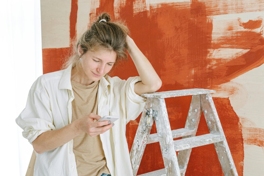 A woman taking a break during home renovation, using her phone near a stepladder and painted wall.