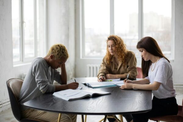Group of focused multiracial classmates taking notes in notebooks while collaborating during preparation for lesson at wooden table with textbook