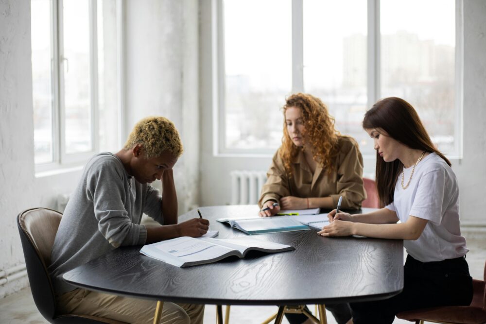 Group of focused multiracial classmates taking notes in notebooks while collaborating during preparation for lesson at wooden table with textbook