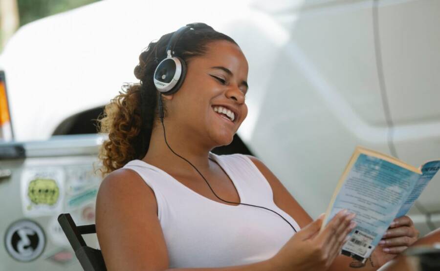 Side view of happy young ethnic female traveler smiling while listening to music in headphones and reading interesting book at campsite on sunny day