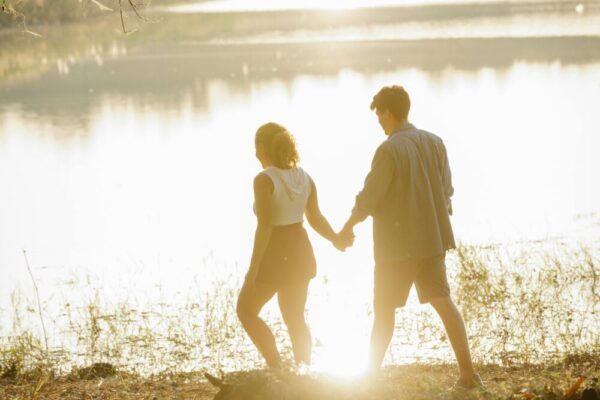 A couple holding hands and walking along a serene lakeside during sunset, capturing a romantic moment outdoors.