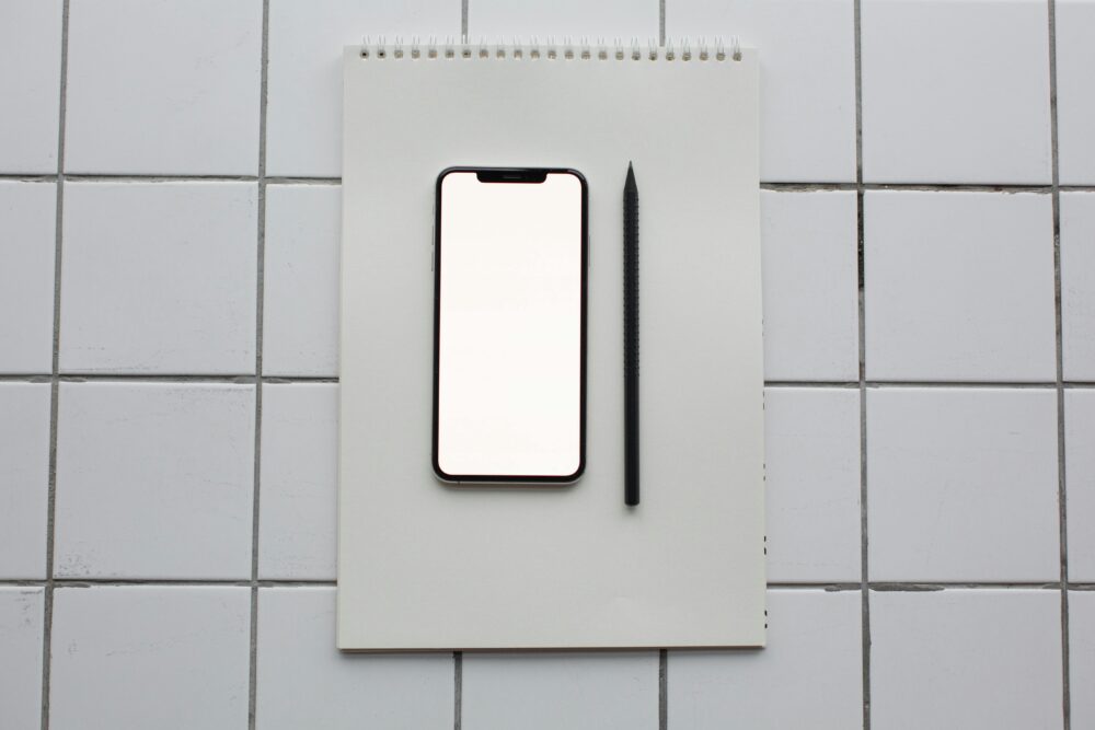Flat lay of a smartphone with blank screen on a notebook next to a pencil, on a tiled surface.