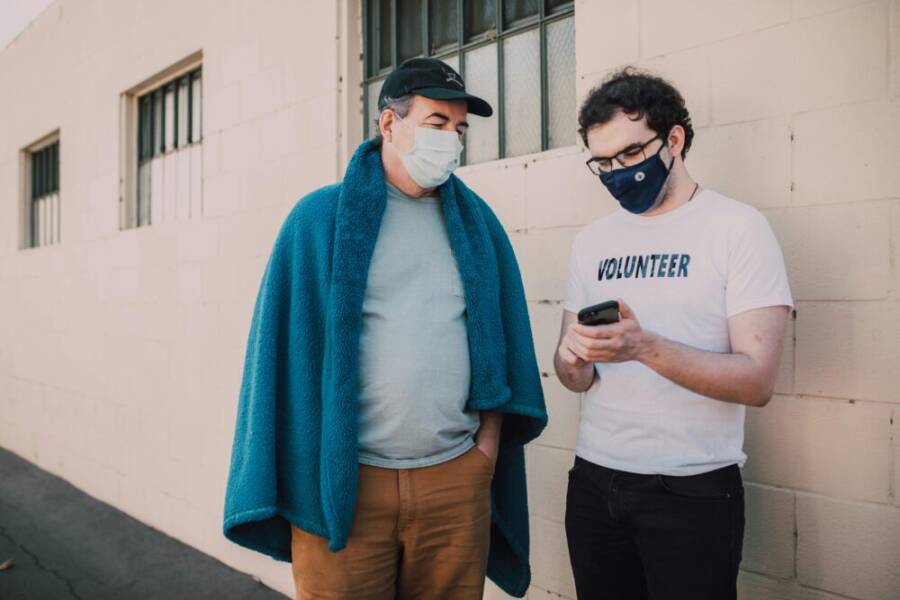 Two masked volunteers stand outside, collaborating on a smartphone project, enhancing community support