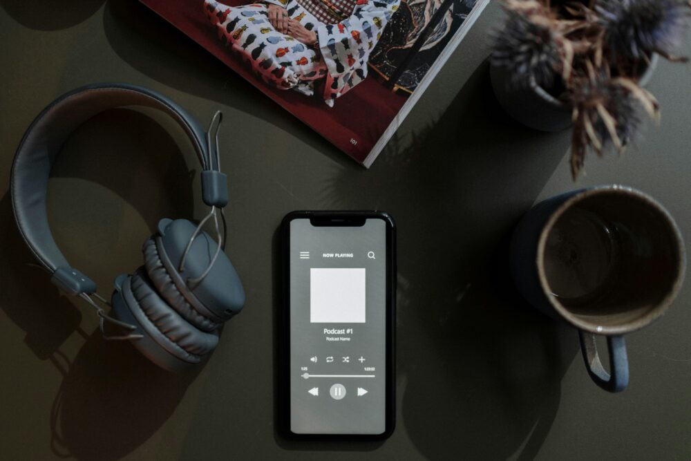 A flat lay image of a smartphone playing a podcast next to headphones and a mug