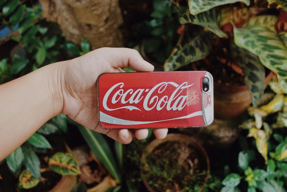 A hand holding a smartphone with a Coca-Cola case against a vibrant garden backdrop