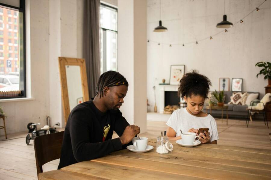 Serious African American father having coffee at table with girl browsing mobile phone in flat