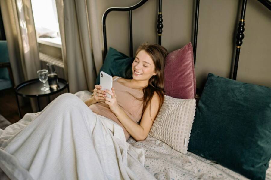 Smiling woman relaxes with smartphone in a cozy, well-lit bedroom setting