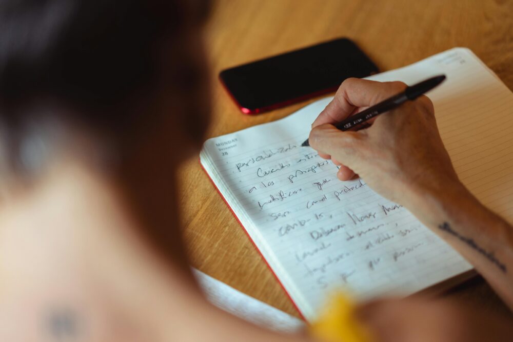 A person writes in a notebook at a wooden desk with a phone nearby