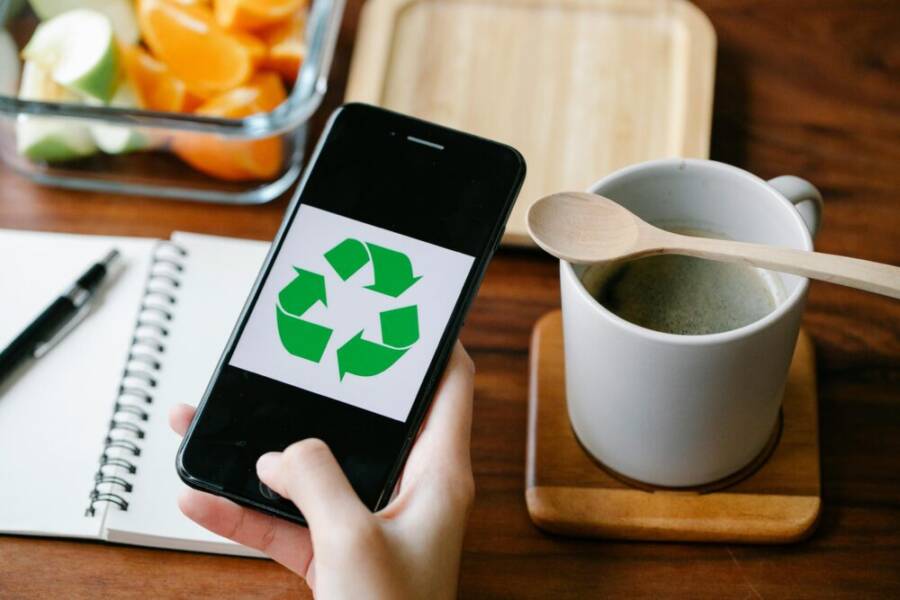 Hand holding smartphone with recycling icon beside coffee and fruit on wooden table