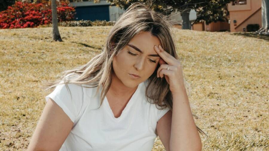 Caucasian woman sitting on grass outdoors with a headache, eyes closed, hand on head.