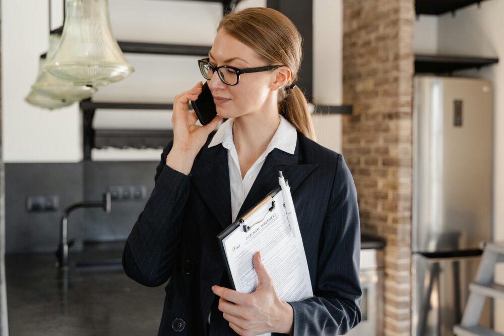 Businesswoman in a black blazer using a cellphone and holding a clipboard in an office setting