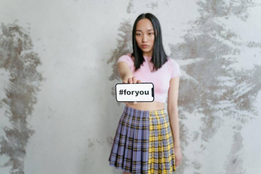 A young woman in a trendy outfit holds a phone displaying #foryou against a textured wall.