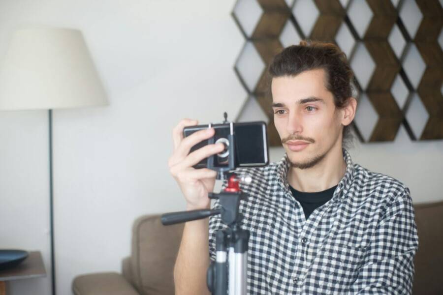 Young man with plaid shirt using a smartphone and tripod to create content indoors in Portugal.