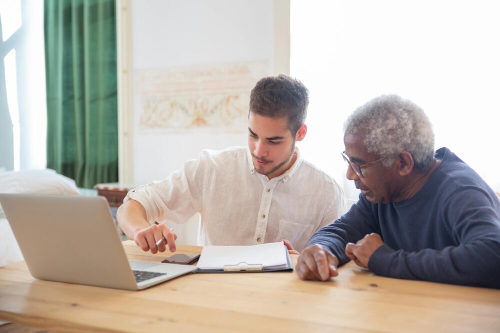 Young man teaches elderly gentleman to use technology at home.