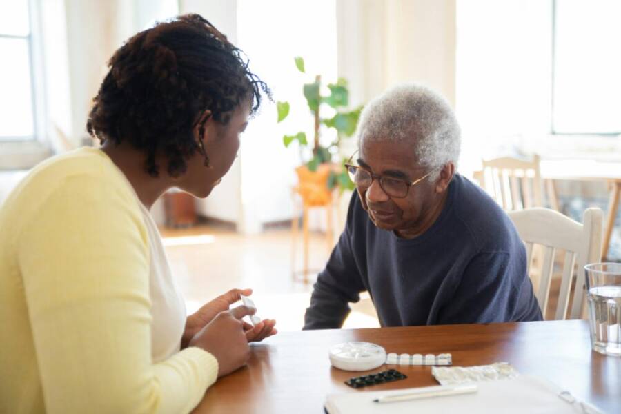 A woman and an elderly man discuss medication at a wooden table in a bright room, highlighting care