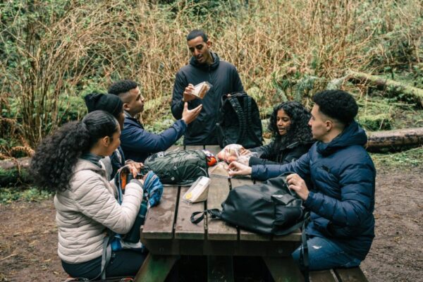 A diverse group of friends sharing snacks at a picnic table in a forest setting during a hiking trip