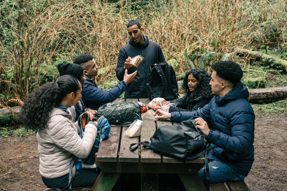 A diverse group of friends sharing snacks at a picnic table in a forest setting during a hiking trip
