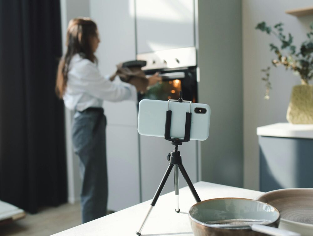 A woman uses a smartphone on a tripod to film her baking process in a modern kitchen.