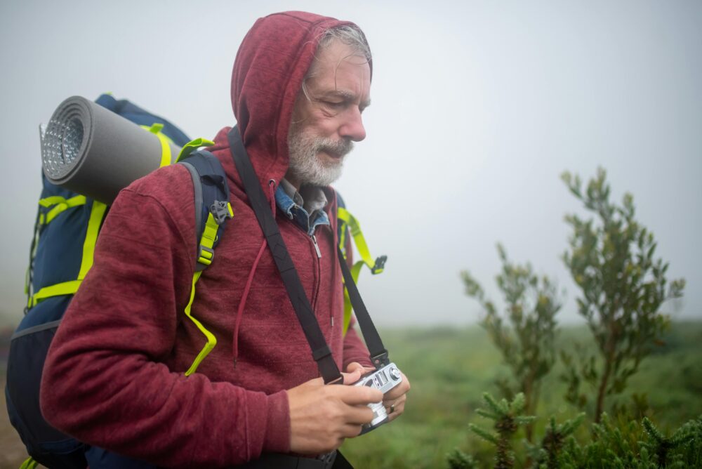 An elderly man hiking through a foggy landscape in Portugal with a backpack and camera.