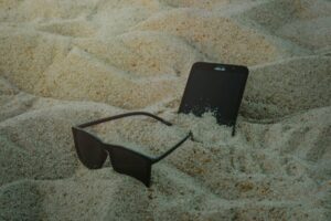 A smartphone and sunglasses partially buried in beach sand, capturing a summer vibe.